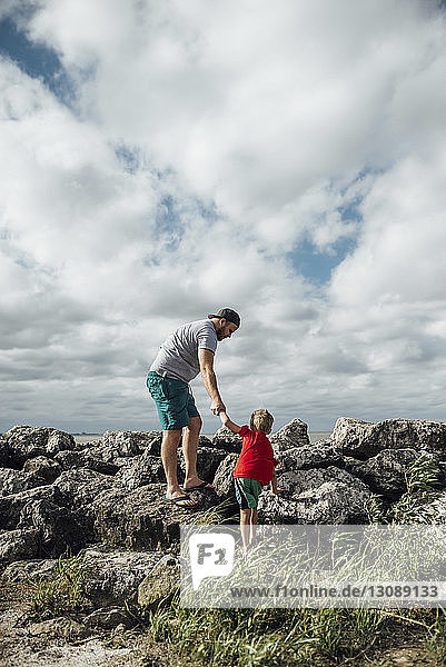 Father holding son's hands while standing on rock at beach against cloudy sky