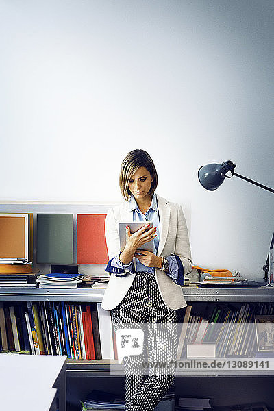 Geschäftsfrau benutzt Tablet-Computer  während sie sich im Büro auf einem Tisch an die Wand lehnt