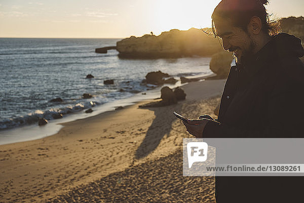 Smiling man using mobile phone while standing at beach against sky during sunset