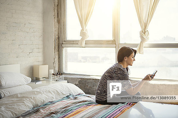 Woman using smart phone while sitting on bed at home