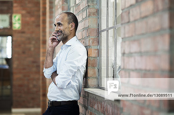 Seitenansicht eines Geschäftsmannes  der mit einem Smartphone spricht  während er an einer Ziegelmauer steht