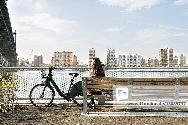 Woman sitting on bench in front of East river against clear sky