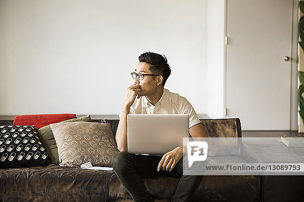 Nachdenklicher Geschäftsmann mit Laptop-Computer  der zu Hause auf dem Sofa sitzt