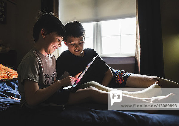 Smiling boy with broken leg using tablet computer while sitting by brother on bed at home