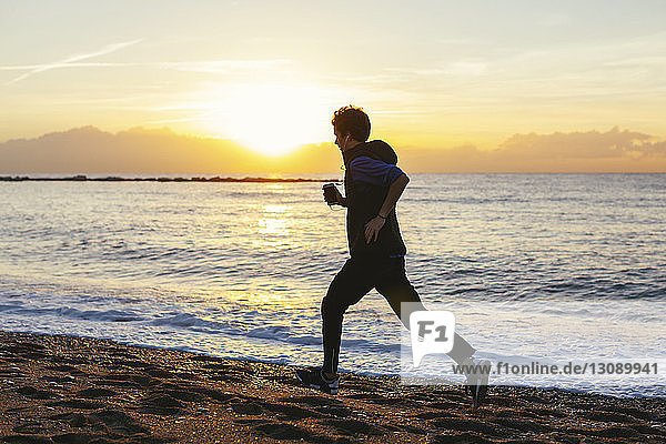 Teenager Junge hört Musik  während er bei Sonnenuntergang am Strand gegen das Meer joggt
