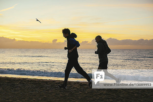 Side view of father and son jogging at beach against cloudy sky during sunset