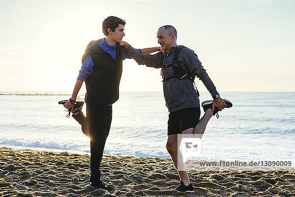 Full length of father and son stretching legs while standing face to face at beach against clear sky during sunset