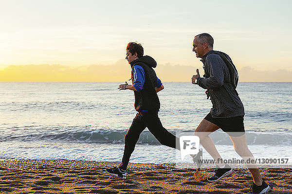 Full length of father and son jogging on shore at beach against sky during sunset