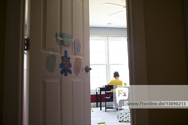 Rear view of boy sitting on chair by table at home seen through doorway