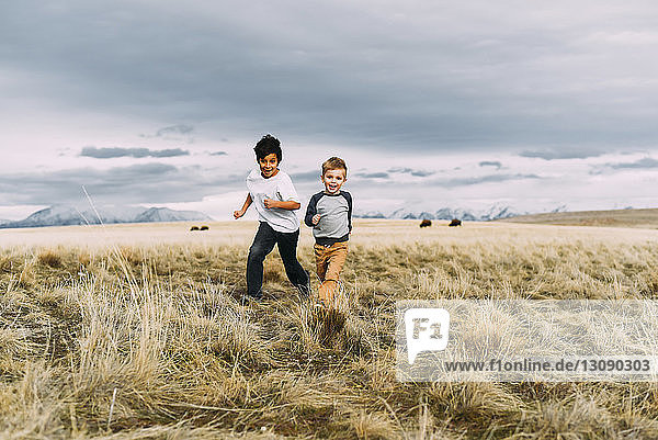 Happy brothers running while playing on grassy field against cloudy sky