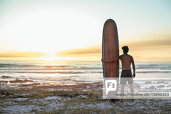 Rear view of shirtless man holding surfboard at beach against clear sky during sunset