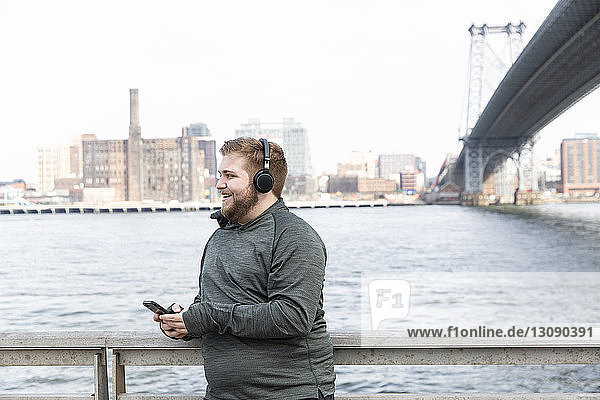 Smiling overweight man listening music on headphones while standing by river against Williamsburg Bridge in city