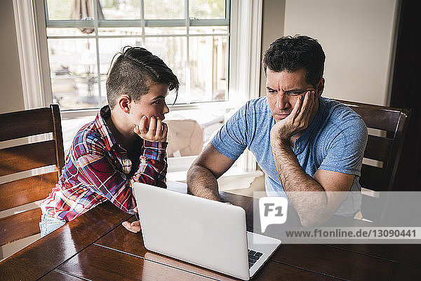 Son looking at stressed father using laptop computer at home