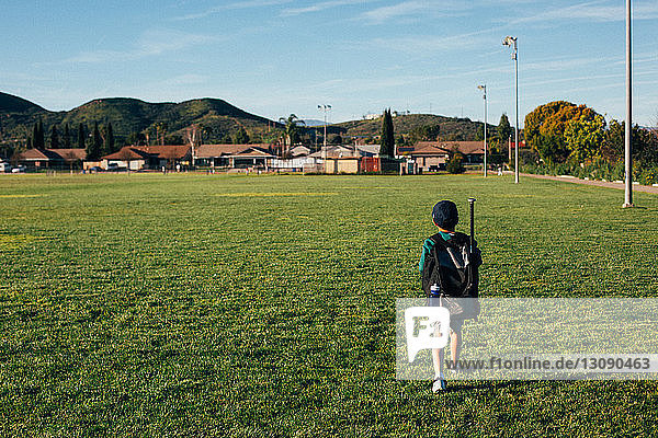 Rear view of boy carrying backpack while walking on grassy field against sky
