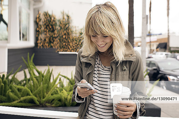 Glückliche junge Frau  die ein Smartphone benutzt  während sie auf der Straße in der Stadt Einwegglas hält