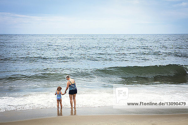 Rückansicht der Enkelin und Großmutter  die am Strand am Ufer stehen und Händchen halten