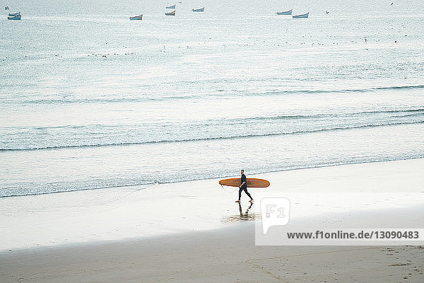 Mann mit Surfbrett in voller Länge beim Strandspaziergang am Ufer