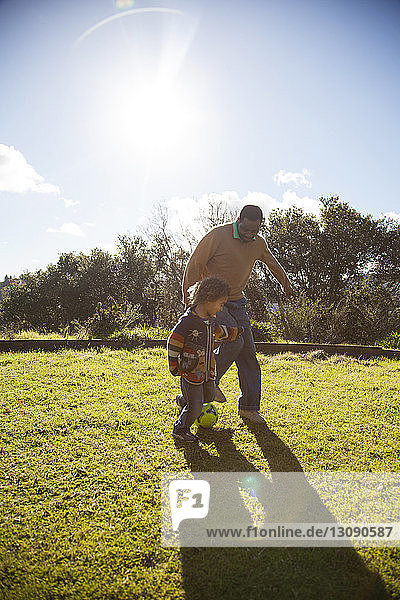 Father and son playing with ball on grassy field against sky during sunny day