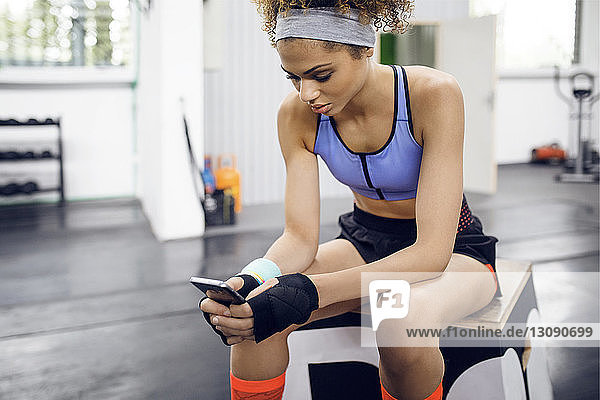 Sportlerin benutzt Smartphone  während sie im Fitnessstudio auf der Box sitzt