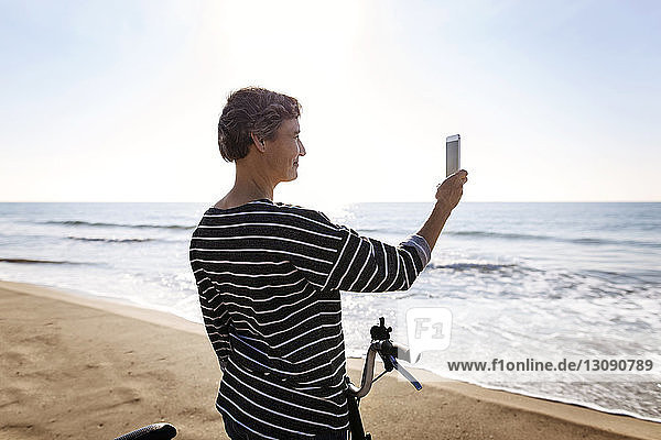 Frau fotografiert mit dem Handy  während sie mit dem Fahrrad am Strand steht