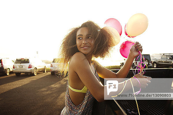 Smiling teenage girl with balloons near car against clear sky