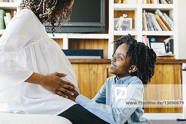 Glückliche Tochter berührt den Bauch der schwangeren Mutter  die zu Hause auf dem Sofa sitzt