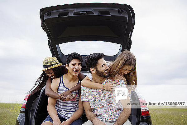 Cheerful friends sitting in car trunk on field against clear sky
