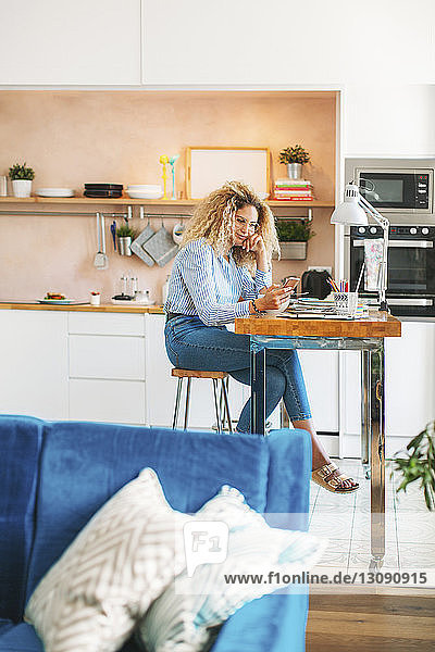 Eine Frau in voller Länge benutzt ein Smartphone  während sie in der Küche am Tisch sitzt