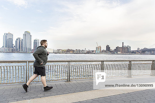 Übergewichtiger Mann in voller Länge joggt auf Brücke am Fluss in der Stadt