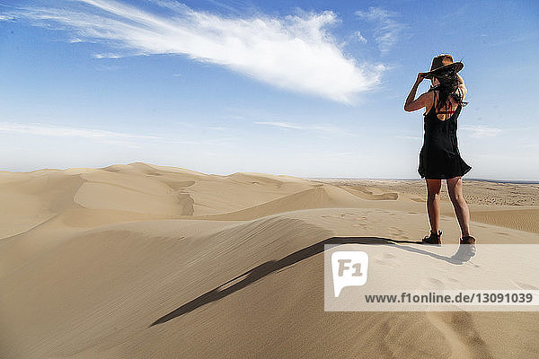 Rear view of young woman wearing hat while walking on sand dune at desert against sky during sunny day