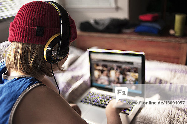 Rückansicht eines Jungen mit Kopfhörern  der seinen Laptop zu Hause am Bett benutzt