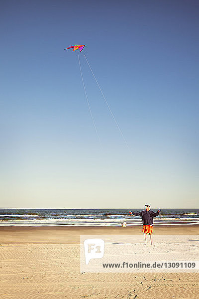 Man flying kite while standing on beach against clear sky