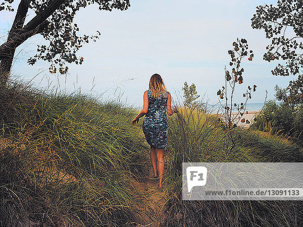 Rear view of woman walking on pathway amidst grassy field against sky