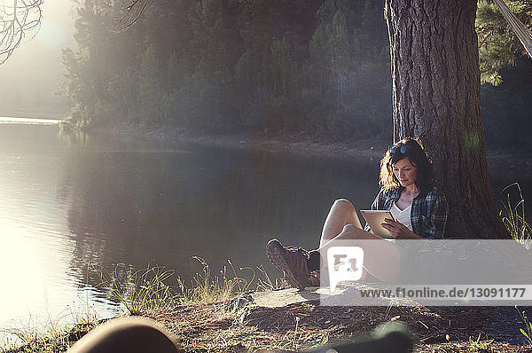 Frau benutzt Tablet-Computer  während sie am Seeufer am Baum sitzt