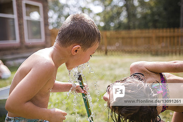 Verspielte Schwester schaut dem Bruder beim Bücken im Hof zu  wie er durch den Gartenschlauch Wasser trinkt