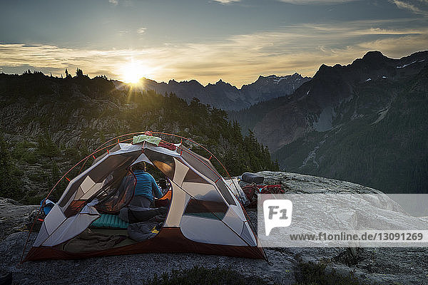 Woman sitting in tent on cliff against mountains during sunset