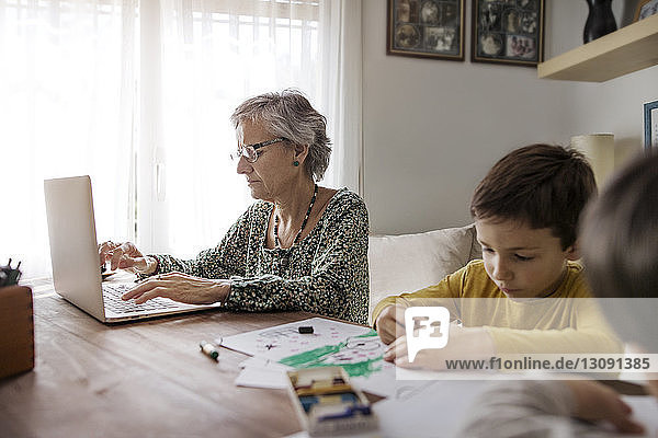 Ältere Frau benutzt Laptop  während Enkel zu Hause Zeichnungen anfertigen