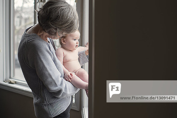 Grandmother holding shirtless granddaughter while standing by window at home
