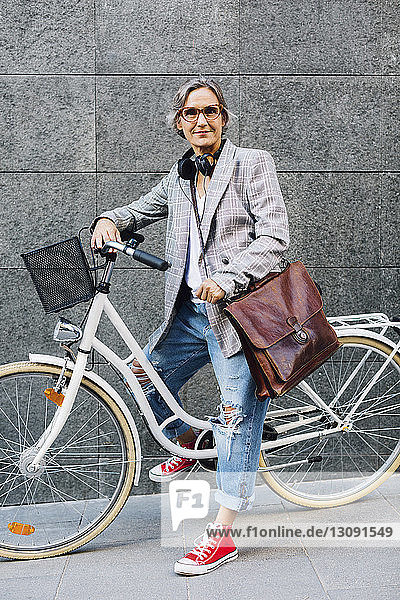 Portrait of confident woman with bicycle against wall on footpath