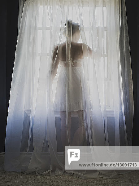 Rückansicht einer Frau  die zu Hause durch ein Fenster durch einen Vorhang schaut