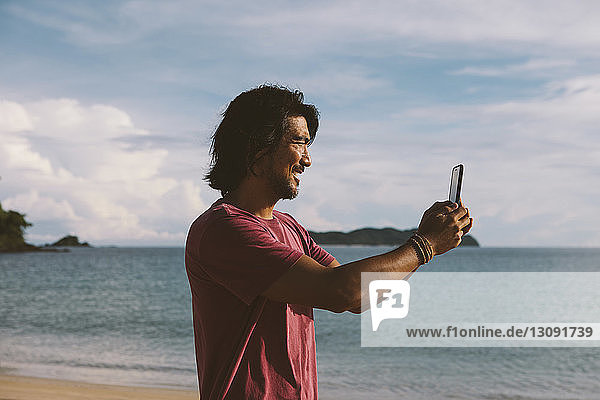 Seitenansicht eines Mannes  der mit einem Mobiltelefon am Strand vor bewölktem Himmel fotografiert