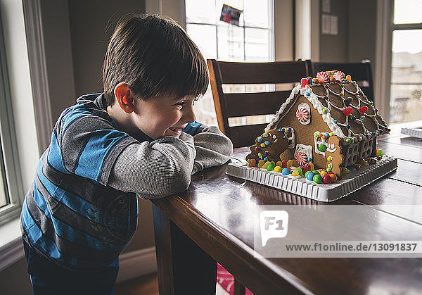 Fröhlicher Junge betrachtet zu Weihnachten Lebkuchenhaus auf Holztisch