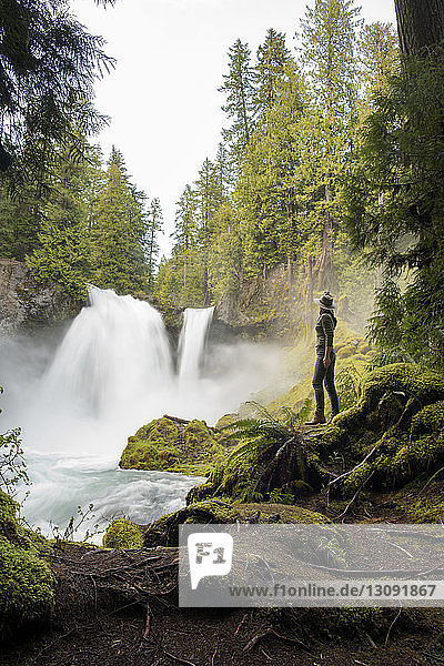 Seitenansicht einer Wanderin  die auf einem moosbewachsenen Felsen stehend den Wasserfall betrachtet