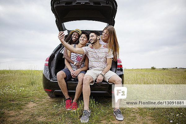 Happy friends taking selfie through smart phone while sitting in car trunk on field against sky