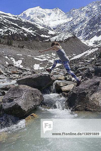 Side view of woman jumping on rocks by river against snowcapped mountains
