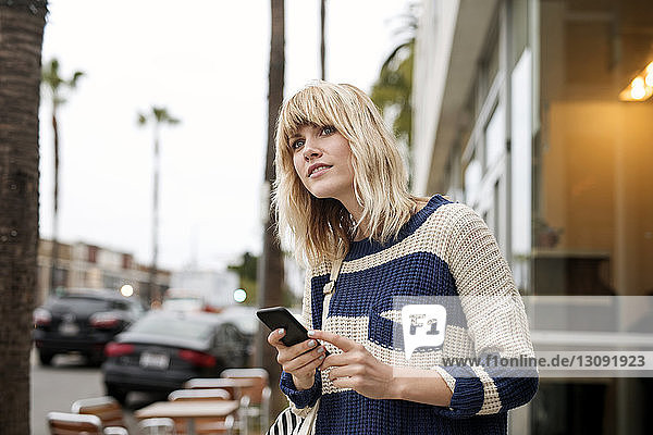 Thoughtful woman holding smart phone by city street