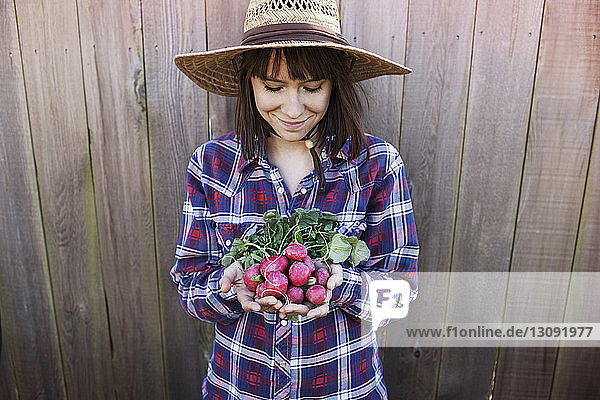 Lächelnde junge Bäuerin hält Radieschen  während sie an einem Holzzaun steht