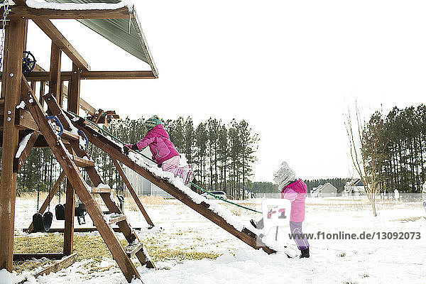 Sisters playing on slide against clear sky during winter