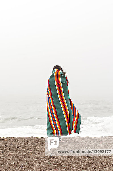 Rückansicht einer in ein Handtuch gehüllten Frau beim Strandspaziergang am Ufer