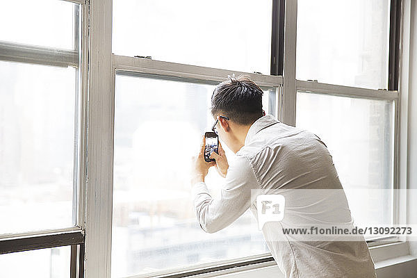 Businessman photographing from window through smart phone in creative office